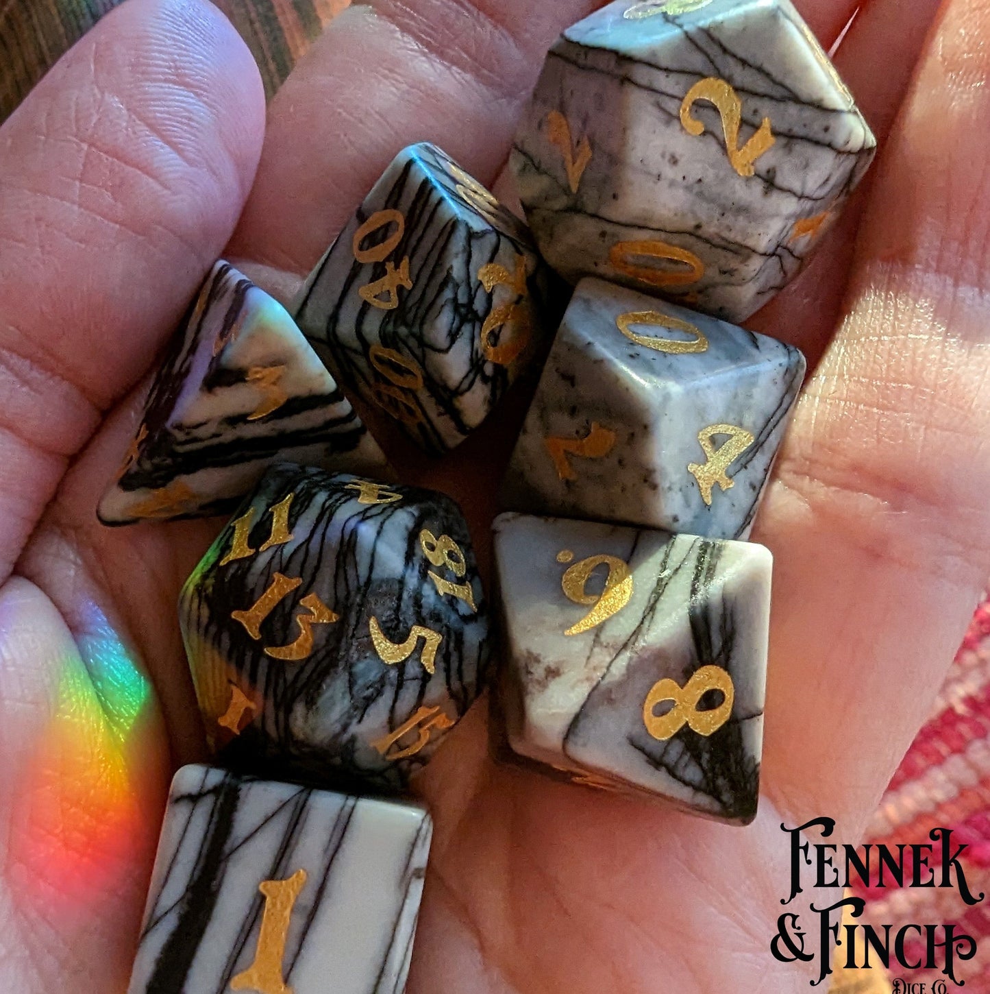 Hand holding Spider Web Jasper gemstone TTRPG dice set with gold numbers, featuring real carved and etched stone dice.