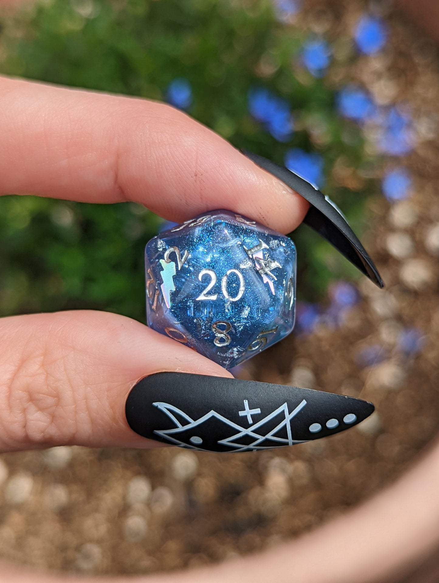 Blue holographic D20 dice from Storm Daddy DnD set held between fingers with black nail art.
