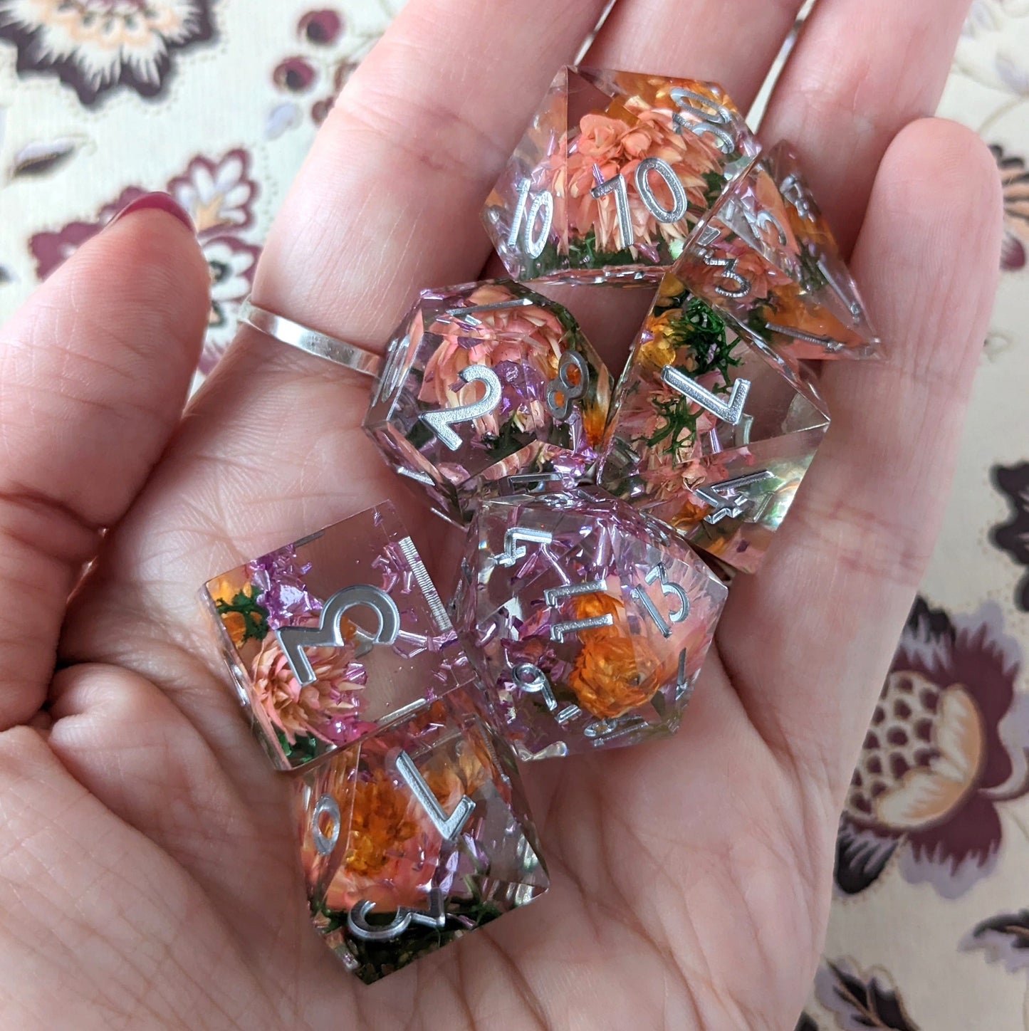 Hand holding Sunny Garden DnD dice set with dried orange and pink flowers inside, featuring sharp edges and standard 7-piece design.
