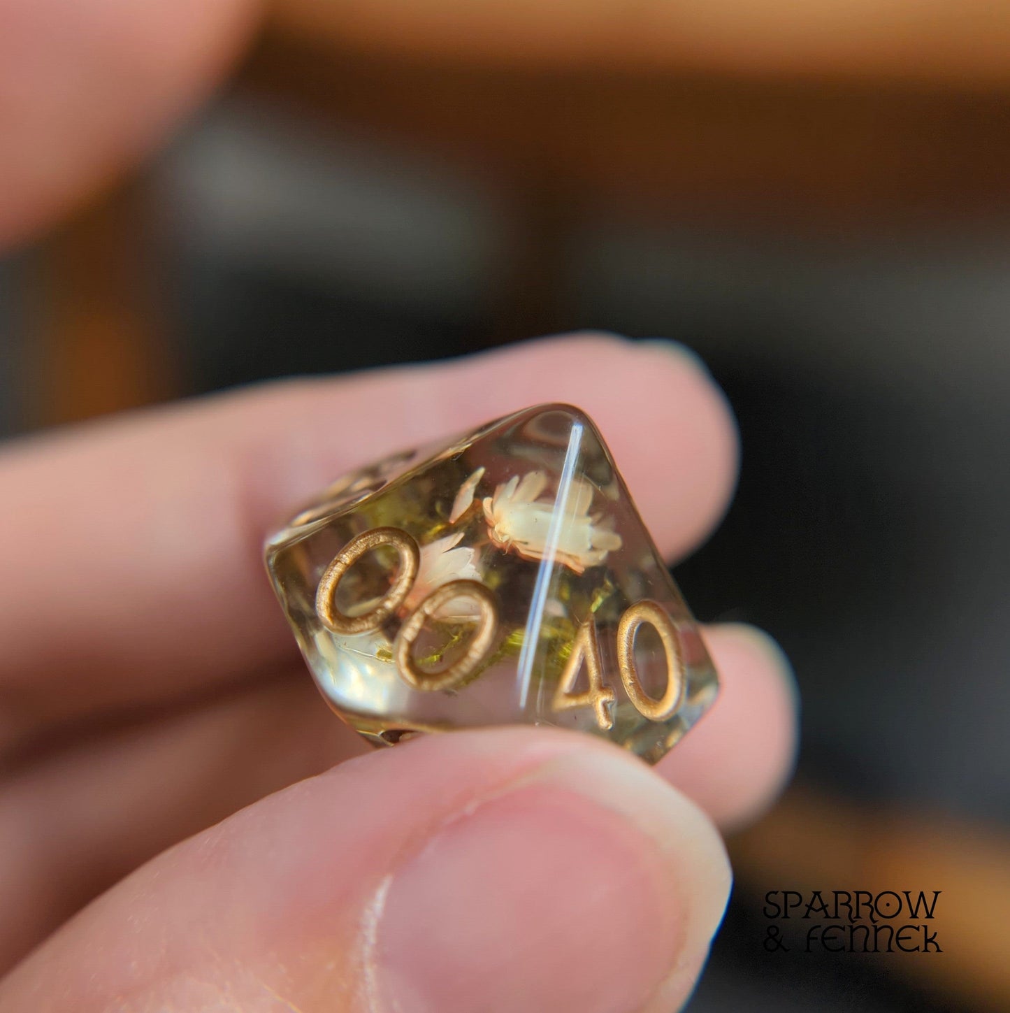 Hand holding resin DnD dice with tiny orange flowers and moss inside, part of a 7-piece polyhedral set for tabletop RPG games.