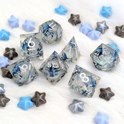 A set of clear acrylic polyhedral dice with blue and black marbled swirls, scattered on a white faux fur background with blue, gray, and brown star-shaped decorative pieces.