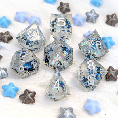A set of clear acrylic polyhedral gaming dice with holographic blue and black swirl patterns, arranged on a white faux fur surface with decorative blue, gray, and brown star-shaped props.