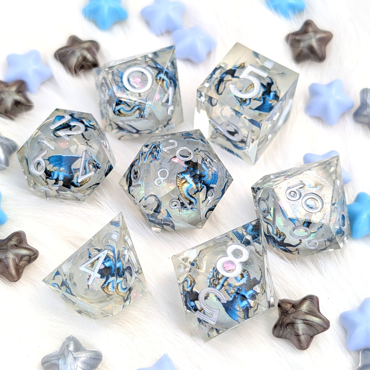 A set of clear acrylic polyhedral dice with blue, black, and white marbled patterns, displayed on a white faux fur background with decorative blue and silver star-shaped props.