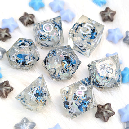 A set of clear acrylic polyhedral dice with blue, black, and white marbled patterns, displayed on a white faux fur background with decorative blue and silver star-shaped props.
