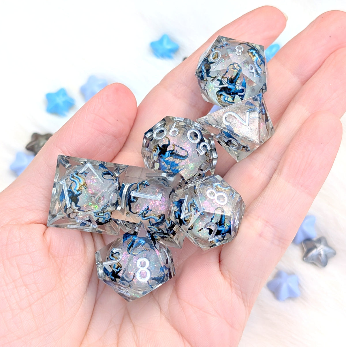 A set of translucent acrylic polyhedral dice with holographic blue, black, and silver swirls, held in a person's hand against a white background with blurred blue star-shaped props.