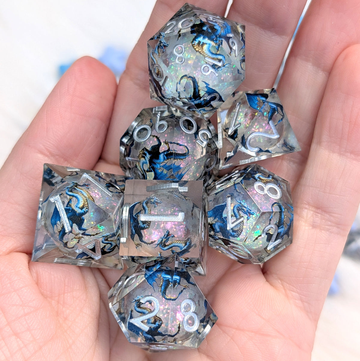 A set of clear acrylic polyhedral gaming dice with blue and black printed graphics and holographic shimmer, held in a person's hand.
