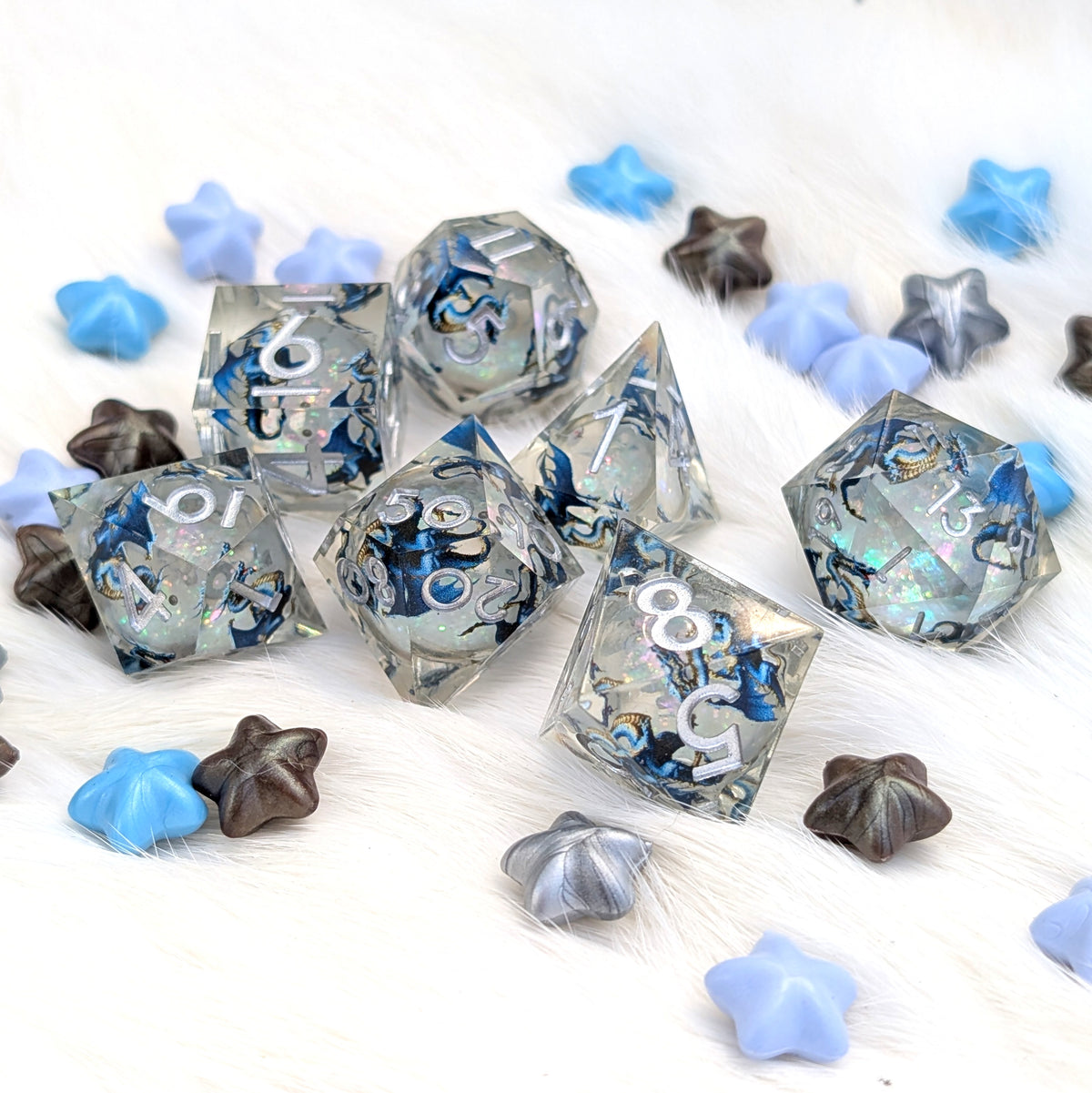 A set of translucent acrylic polyhedral dice with blue and white swirl patterns, scattered on a white faux fur surface alongside blue, gray, and brown star-shaped beads.