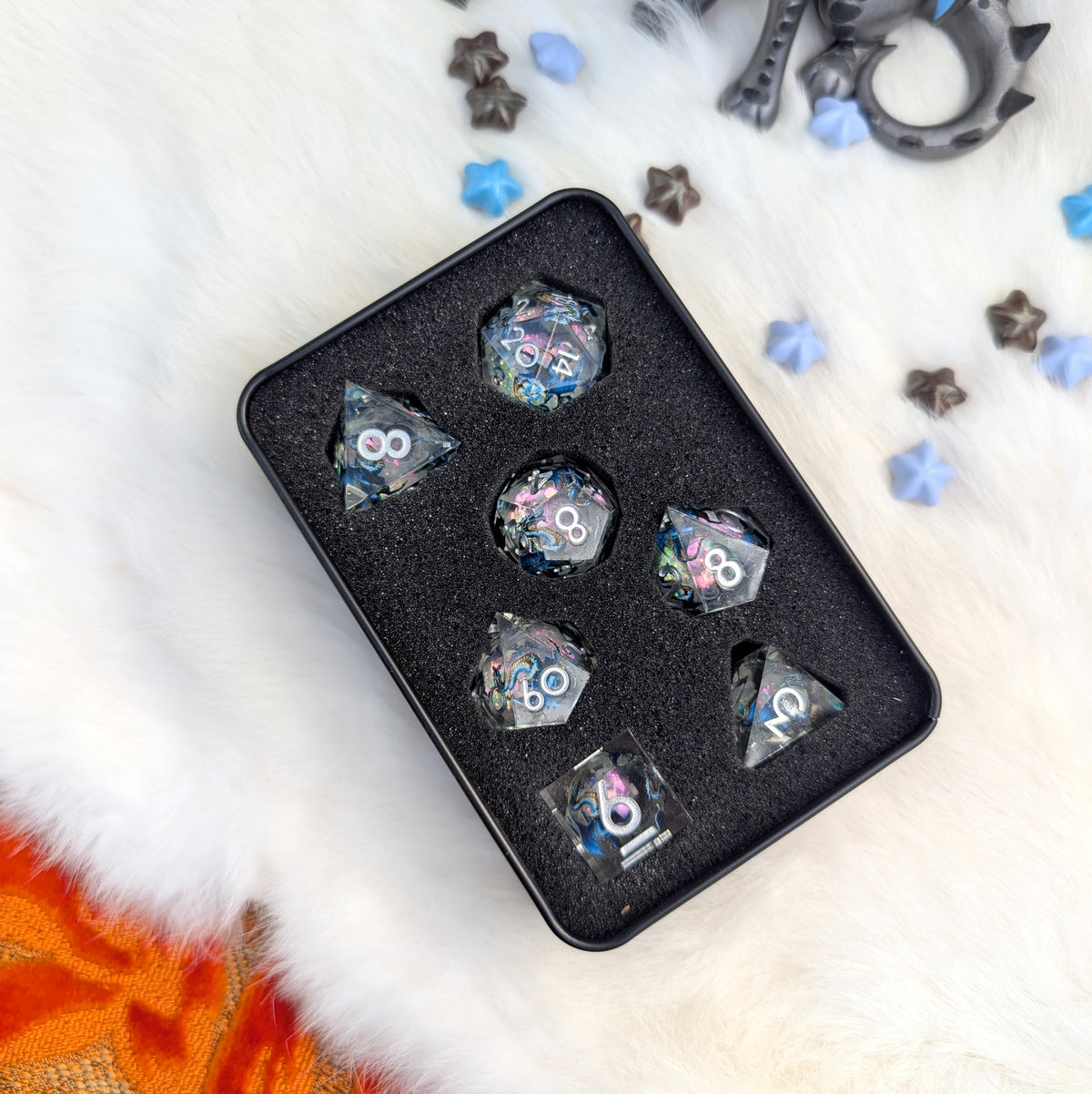 A set of holographic polyhedral gaming dice displayed in a black foam tray inside a black metal tin, placed on a white faux fur surface with scattered star-shaped accessories.