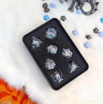 A set of holographic polyhedral gaming dice displayed in a black foam tray inside a black metal tin, placed on a white faux fur surface with scattered star-shaped accessories.