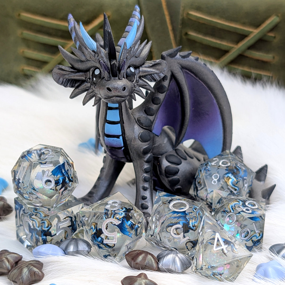A fantasy dragon figurine in dark gray and black with bright blue accents, posed among clear polyhedral gaming dice and shell-like beads on a white faux-fur surface.
