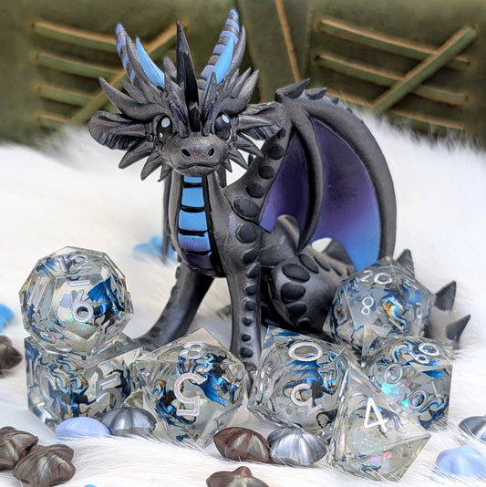 A fantasy dragon figurine in dark gray and black with bright blue accents, posed among clear polyhedral gaming dice and shell-like beads on a white faux-fur surface.
