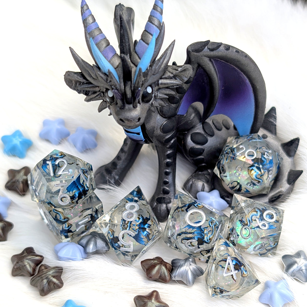 A fantasy dragon figurine in dark gray and black with blue and purple accents, posed among clear polyhedral dice and star-shaped tokens on a white furry surface.