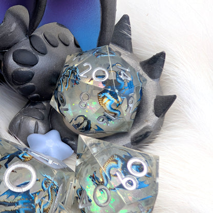 A set of translucent acrylic gaming dice with blue and gold dragon illustrations and holographic shimmer, displayed on a white faux fur surface next to a black plush toy.