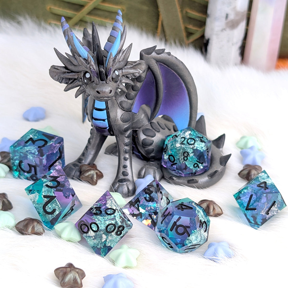 A fantasy dragon figurine in dark gray with blue and purple accents, posed among translucent blue and purple gaming dice on a white faux-fur surface with small decorative shells.