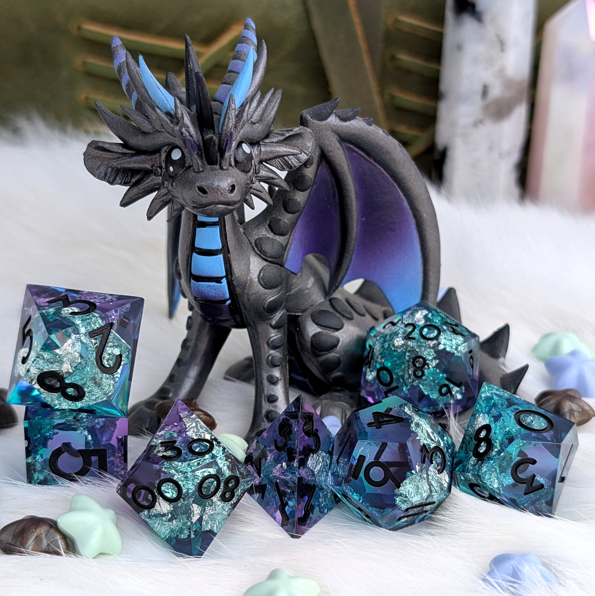 A fantasy dragon figurine in dark gray with bright blue accents, posed among matching translucent purple and blue polyhedral dice on a white faux-fur surface with blurred decorative background elements.