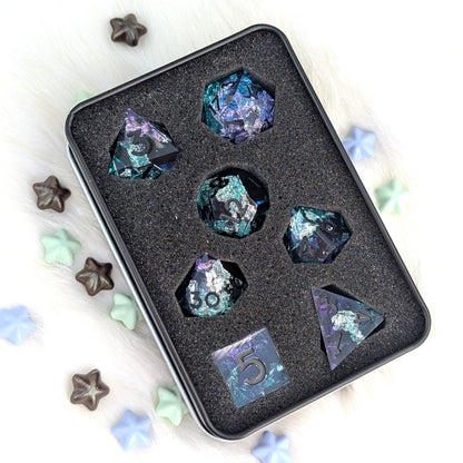 A set of holographic iridescent polyhedral gaming dice displayed in a black foam tray inside a square metal tin, with decorative star and heart-shaped candies on a white furry surface.