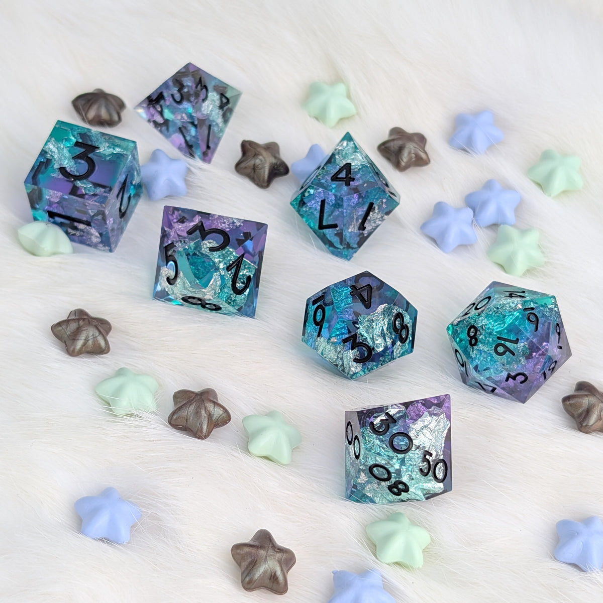 A set of translucent polyhedral gaming dice with a glittery teal, blue, and purple design, arranged on a white faux fur surface with decorative pastel star and shell-shaped props.
