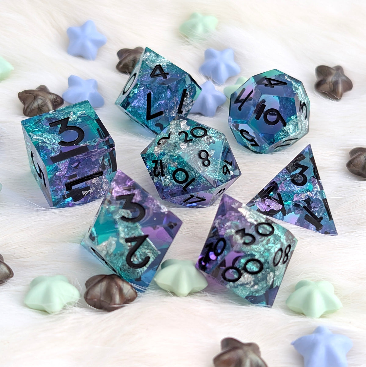 A set of translucent polyhedral gaming dice with a blue, purple, and teal glitter pattern, displayed on a white faux fur surface with decorative star and shell-shaped candies.