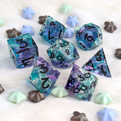 A set of translucent polyhedral gaming dice with a blue, purple, and teal glitter pattern, displayed on a white faux fur surface with decorative star and shell-shaped candies.