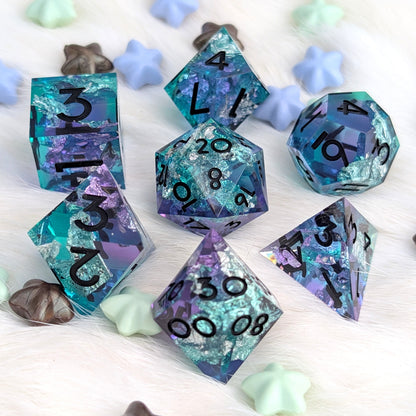 A set of polyhedral gaming dice with a glittery teal, blue, and purple marbled pattern and black printed numbers, arranged on a white faux fur surface with small decorative star and shell props.