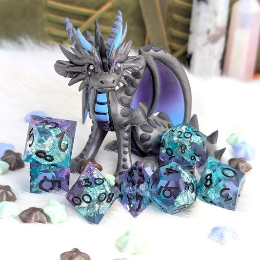A fantasy dragon figurine in dark gray with blue accents, surrounded by matching translucent polyhedral dice in purple, teal, and blue with black numbering, arranged on a white furry surface with small decorative shells and blurred background objects.