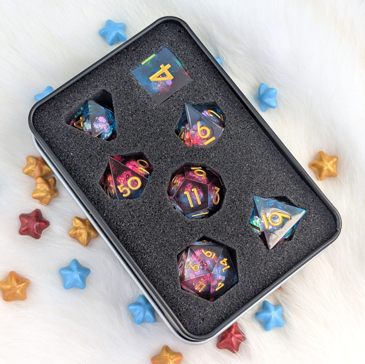 A set of multicolor holographic polyhedral dice displayed in a black foam-lined metal tin, surrounded by scattered star-shaped confetti on a white faux fur background.