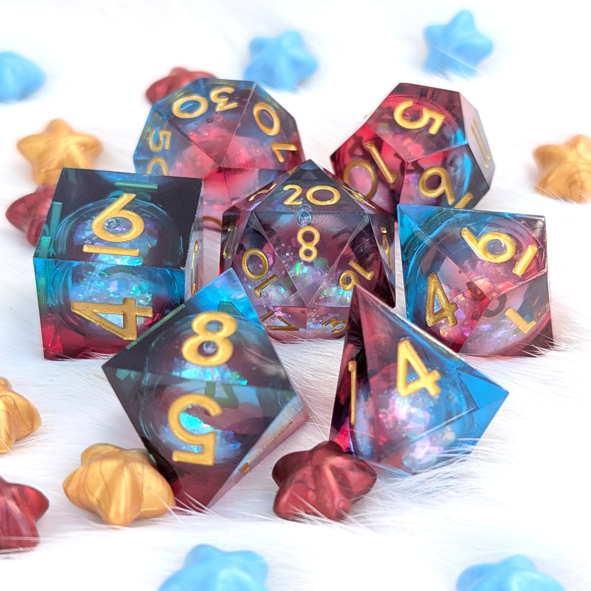 A set of translucent polyhedral gaming dice with gold numbers, arranged on a white furry surface with scattered star-shaped and shell-shaped decorative props. The dice show a galaxy-like color shift between blue, purple, pink, and red.