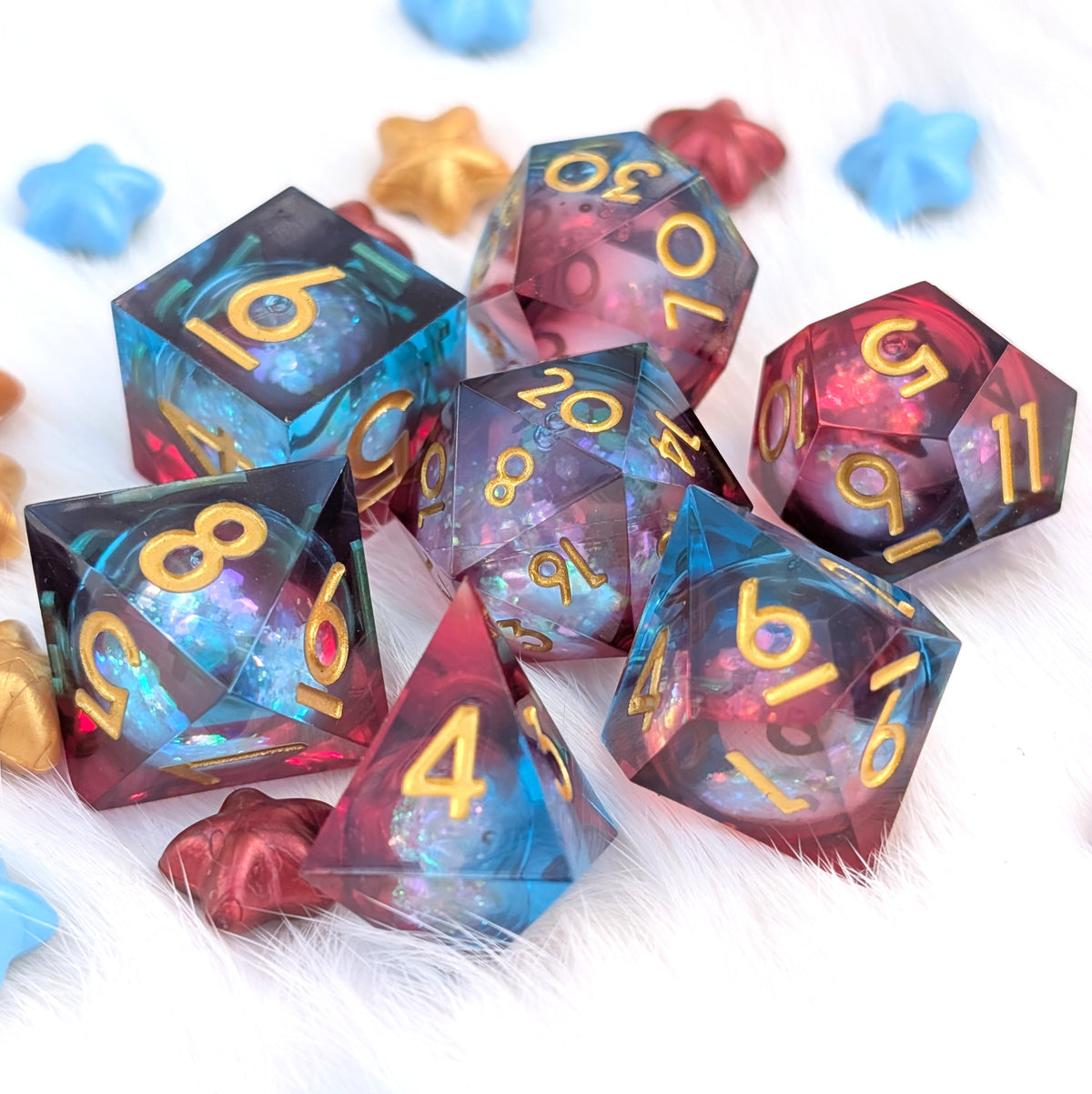 A set of translucent polyhedral dice with a galaxy-like blue, purple, and pink internal pattern and gold numbers, arranged on a white furry surface with small star-shaped confetti.