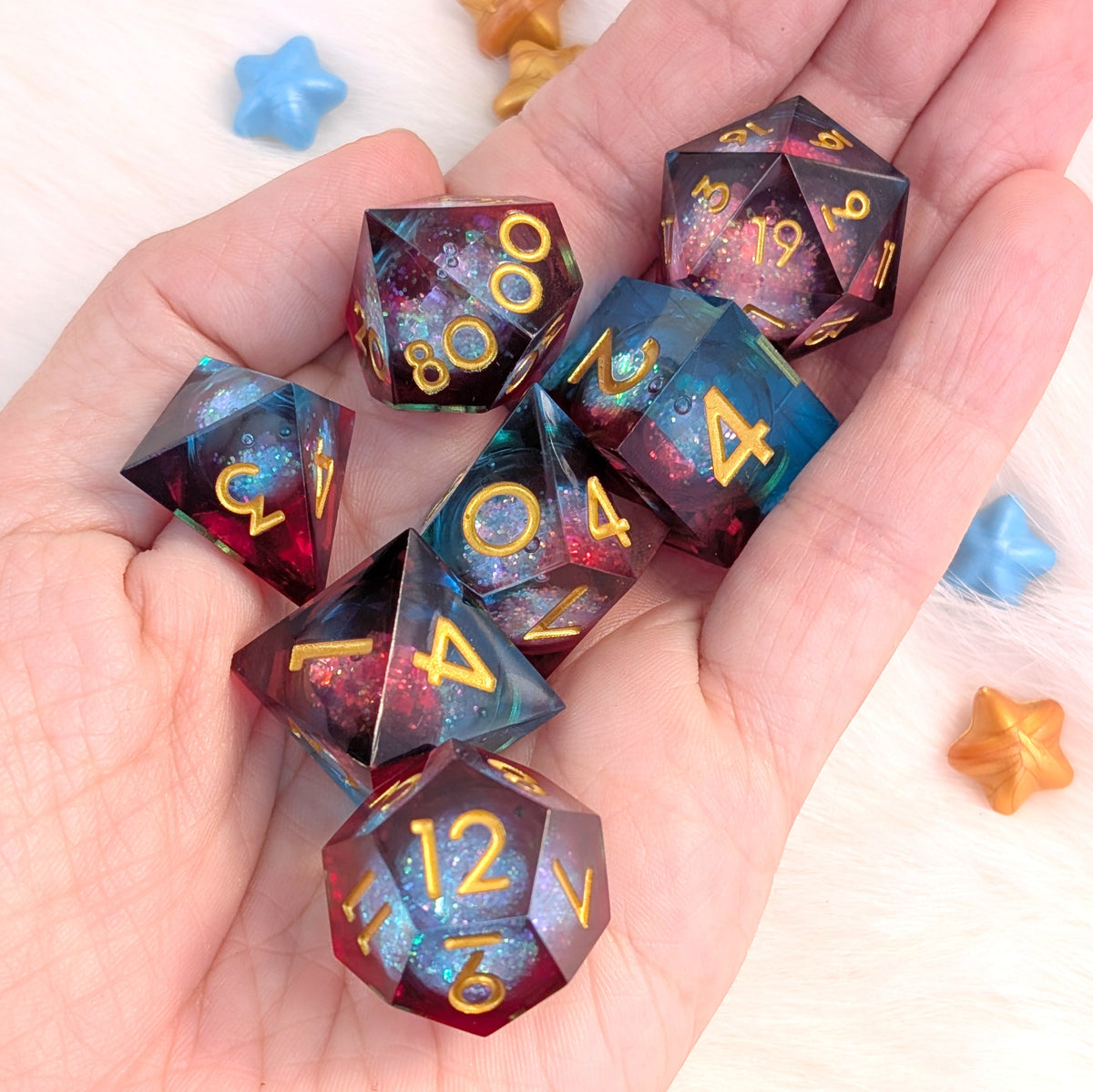 A set of translucent polyhedral dice with gold numbers and a galaxy nebula design, held in a person's hand. The dice feature a gradient of deep purple, red, and blue with sparkling cosmic details.