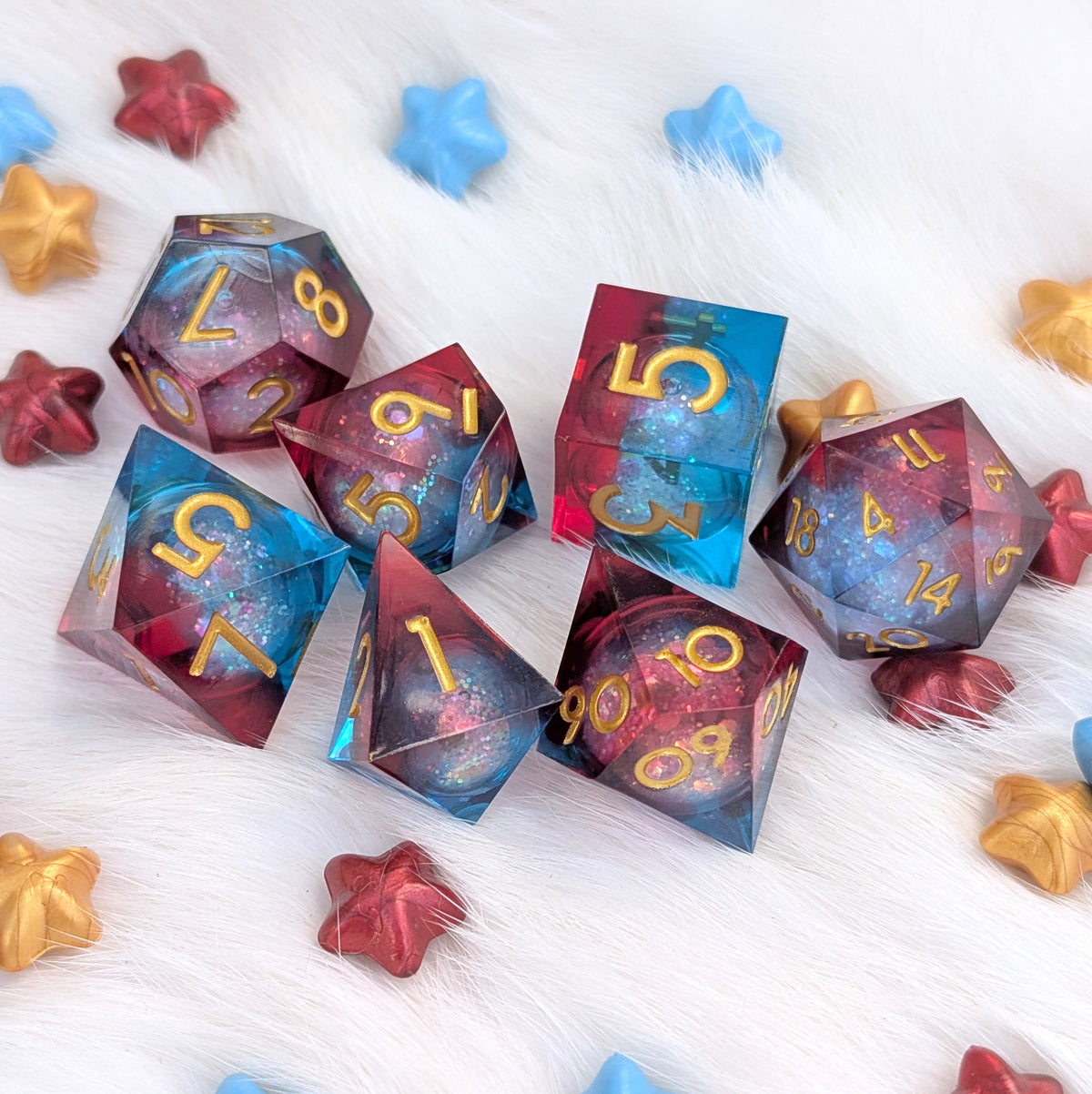 A set of translucent polyhedral gaming dice with a blue, purple, and pink galaxy glitter design and gold numbers, arranged on a white faux fur surface with scattered star-shaped decorative pieces.