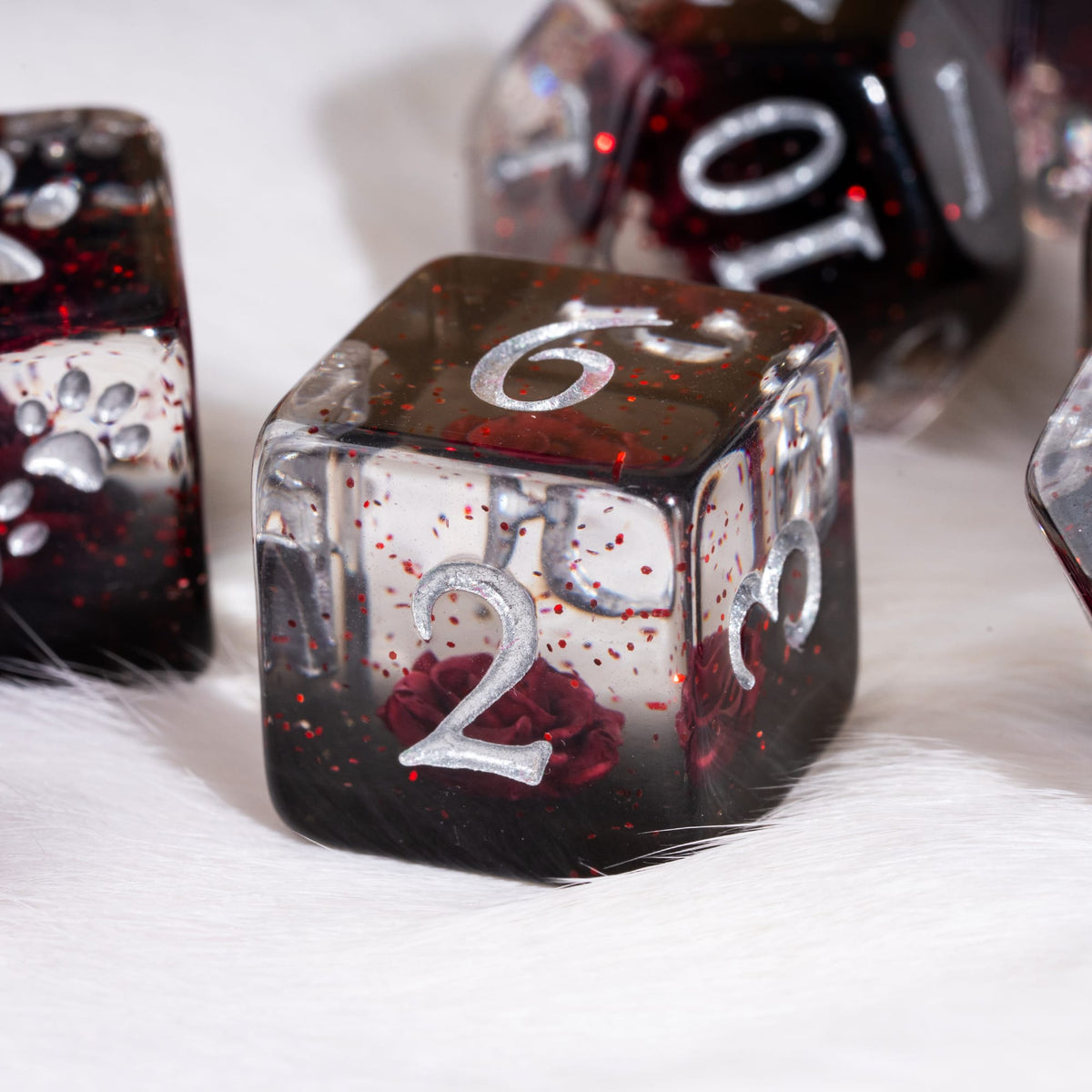 A close-up of a translucent polyhedral die set resting on white faux fur. The main visible die is a d6 with a silver engraved number 2, showing a dark red and black glittery resin body with embedded red rose details.