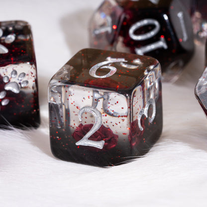 A close-up of a translucent polyhedral die set resting on white faux fur. The main visible die is a d6 with a silver engraved number 2, showing a dark red and black glittery resin body with embedded red rose details.