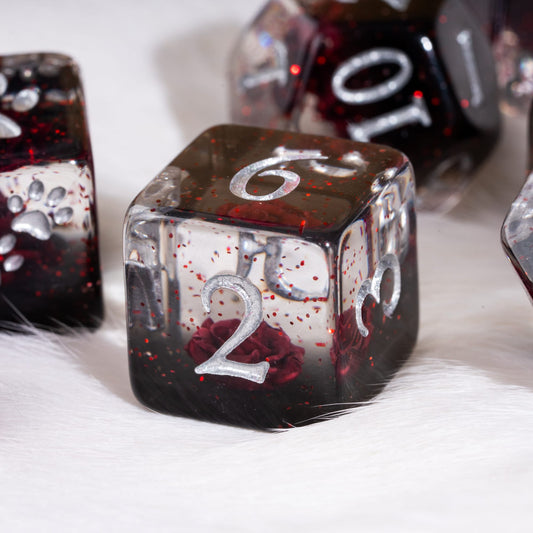 A close-up of a translucent polyhedral die set resting on white faux fur. The main visible die is a d6 with a silver engraved number 2, showing a dark red and black glittery resin body with embedded red rose details.