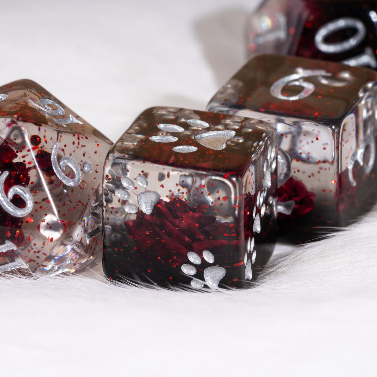 A close-up of clear polyhedral dice filled with dark red glitter and silver paw-print designs, resting on a white furry surface.