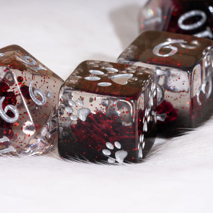 A close-up of clear polyhedral dice filled with dark red glitter and silver paw-print designs, resting on a white furry surface.