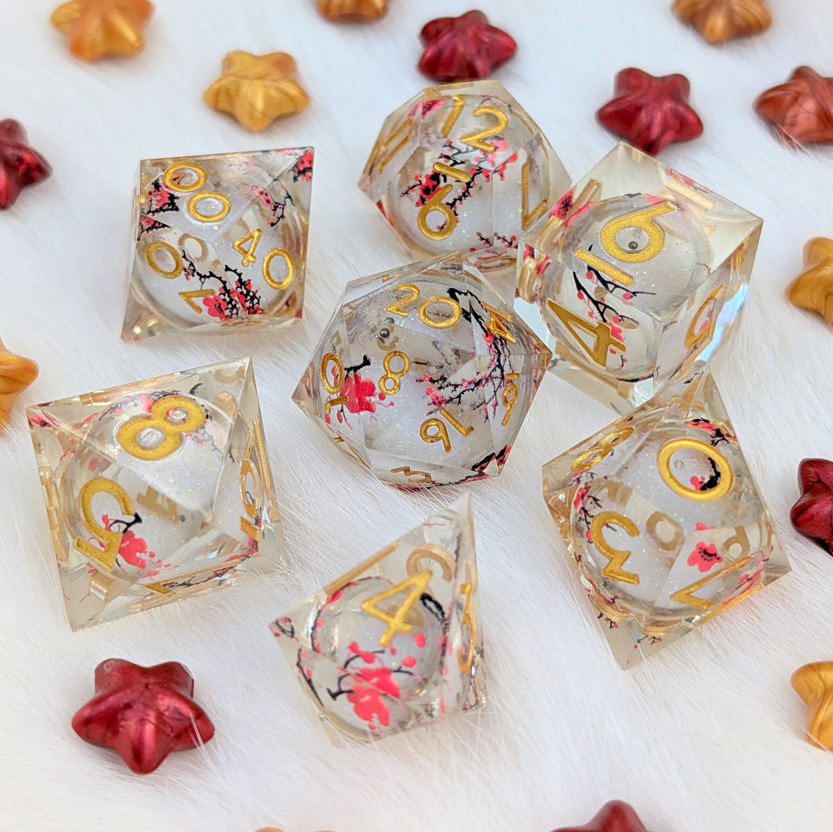 A set of clear acrylic polyhedral dice with gold numbers and pink cherry blossom floral details, arranged on a white faux fur surface with small decorative star-shaped props.