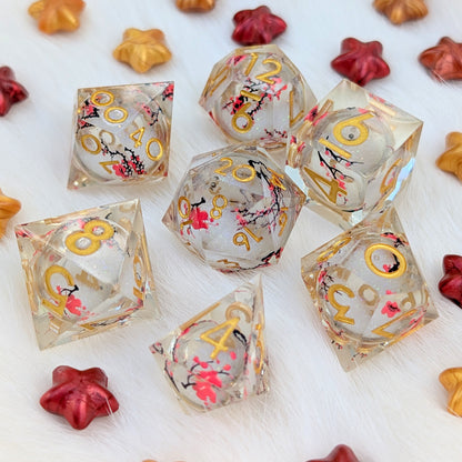 A set of clear acrylic polyhedral dice with gold numbers and pink cherry blossom floral details, arranged on a white faux fur surface with small decorative star-shaped props.