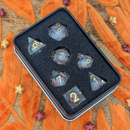 A 7-piece polyhedral tabletop gaming dice set in a black metal tin with a black foam insert, photographed from above on an orange patterned fabric background with small star-shaped props.