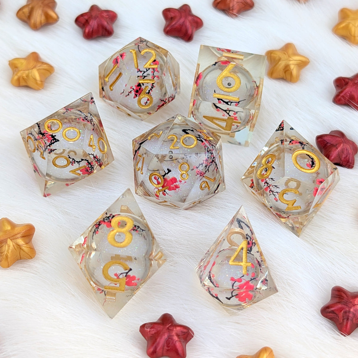 A set of transparent acrylic polyhedral dice with gold numbers and pink cherry blossom floral designs, arranged on a white faux fur background with scattered red and gold star-shaped decorative props.