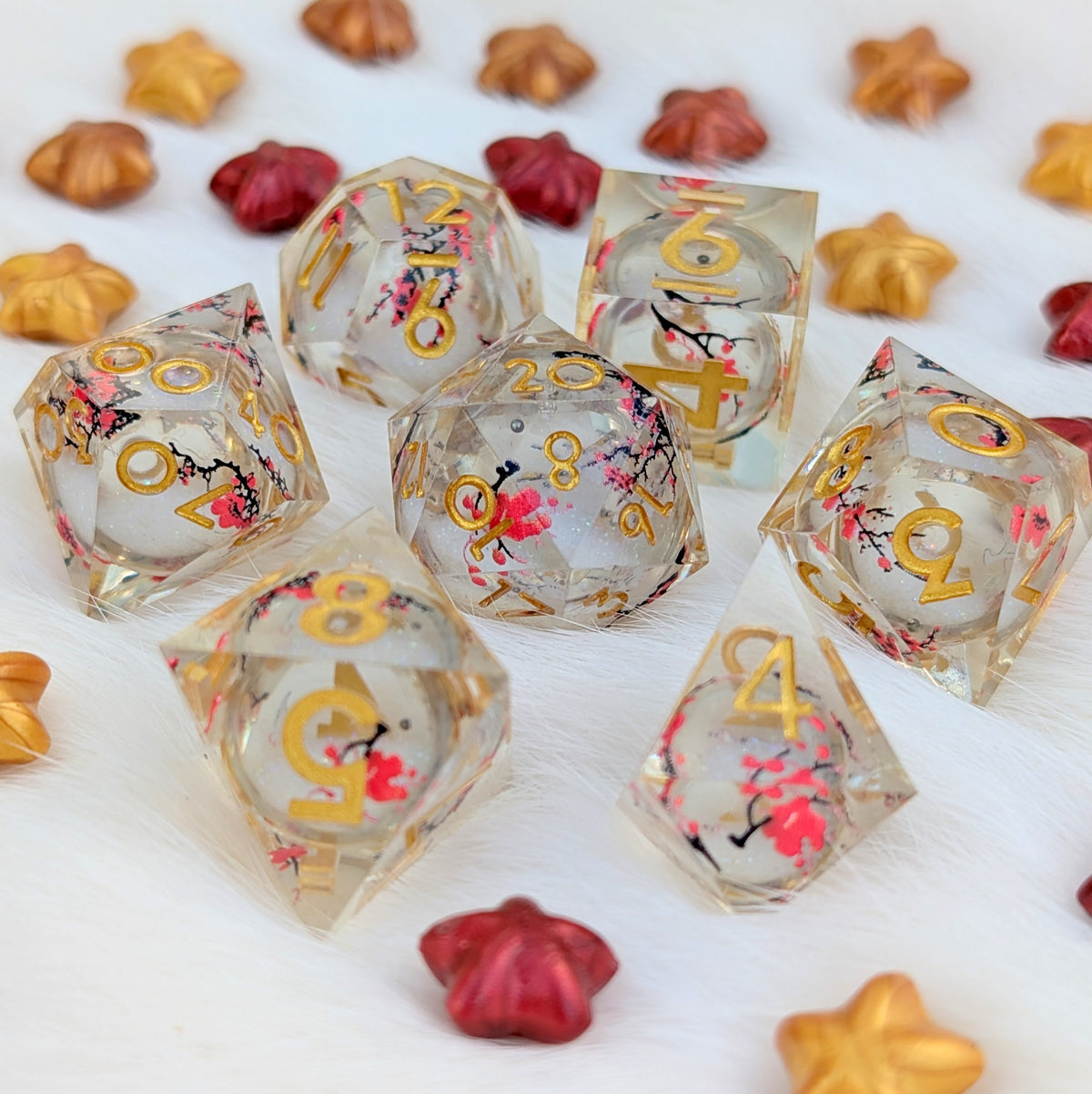 A set of clear acrylic d20 dice with gold numbers and pink cherry blossom floral designs, displayed on a white faux fur surface with scattered decorative star and flower pieces.