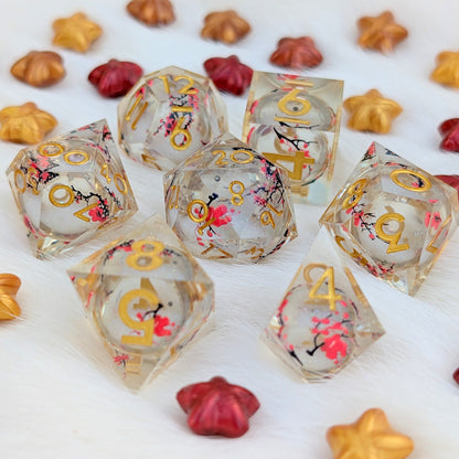 A set of clear acrylic d20 dice with gold numbers and pink cherry blossom floral designs, displayed on a white faux fur surface with scattered decorative star and flower pieces.