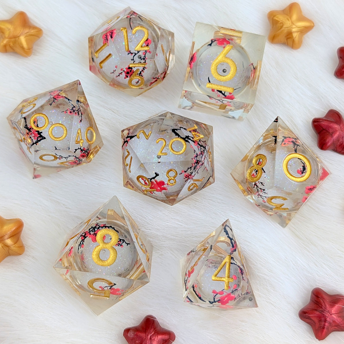A set of transparent acrylic polyhedral dice with gold-colored numbers and pink cherry blossom floral designs, arranged on a white textured background with decorative gold and red star cutouts.