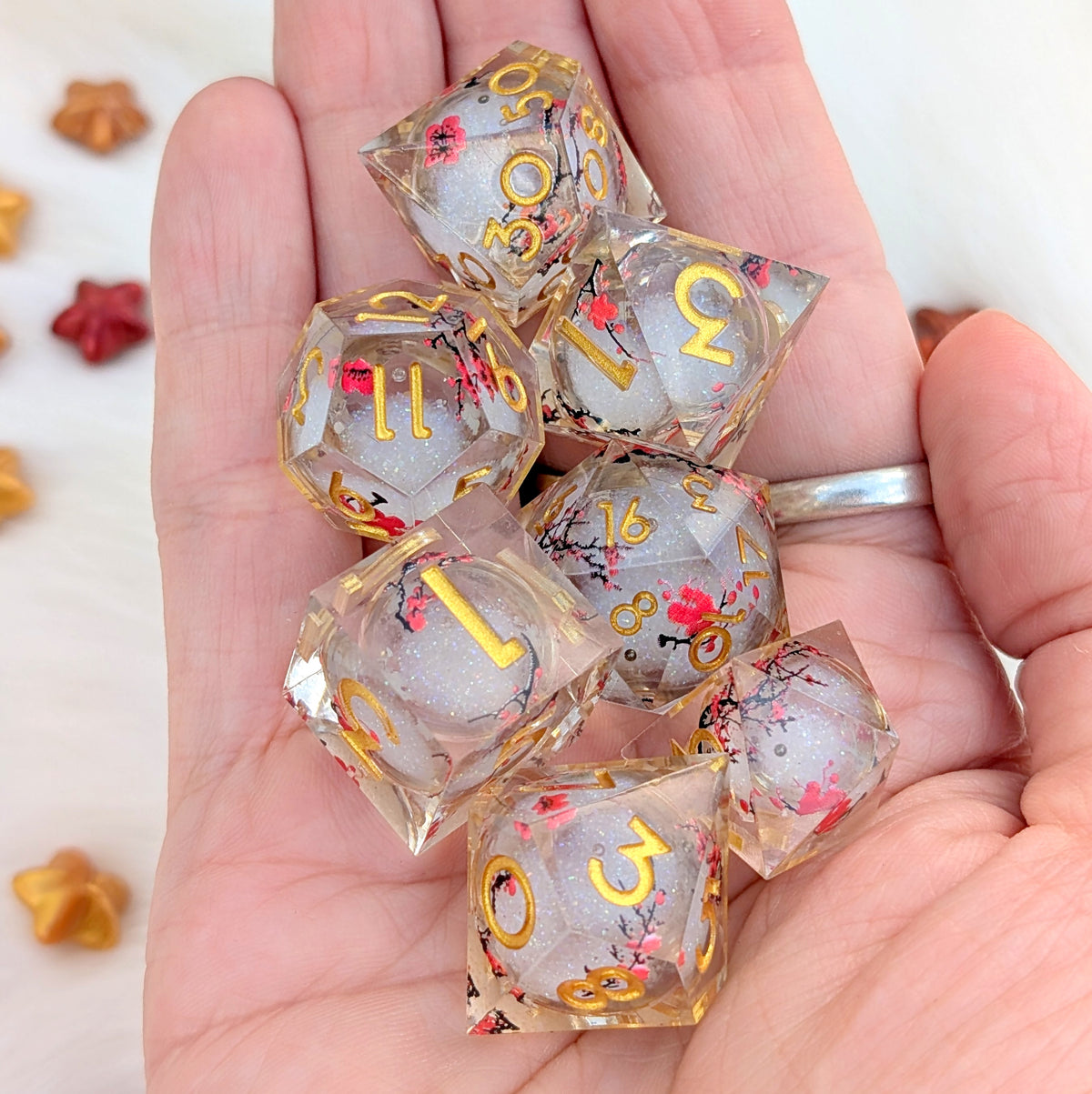 A set of transparent resin polyhedral dice with gold numbers and red floral details, held in a person's hand.