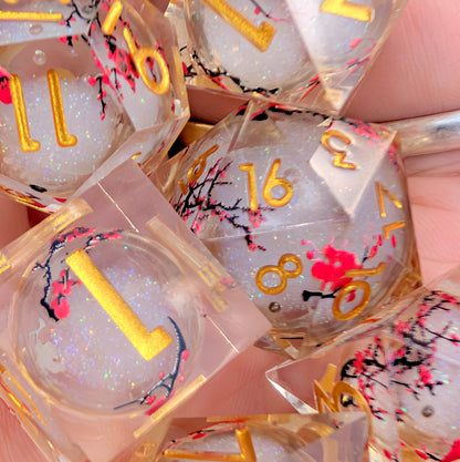A close-up of clear acrylic polyhedral dice filled with white glitter, decorated with gold numbers, black cherry blossom branches, and pink flowers, held in a hand.