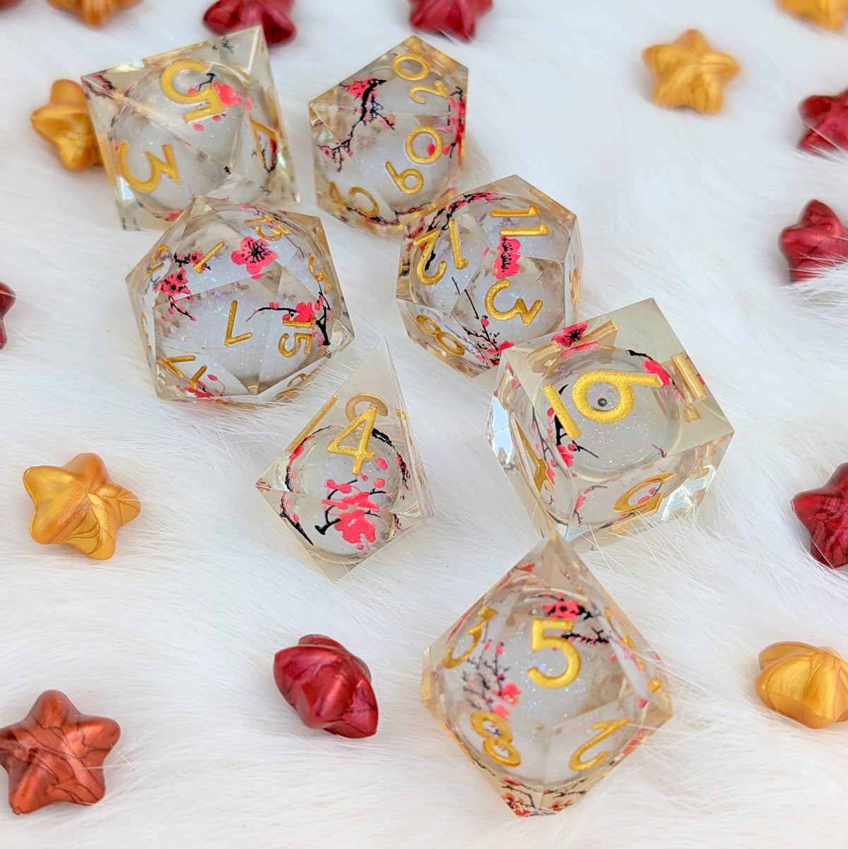 A set of clear acrylic polyhedral gaming dice with gold numbers and pink cherry blossom floral designs, arranged on a white faux fur surface with decorative star-shaped candies.