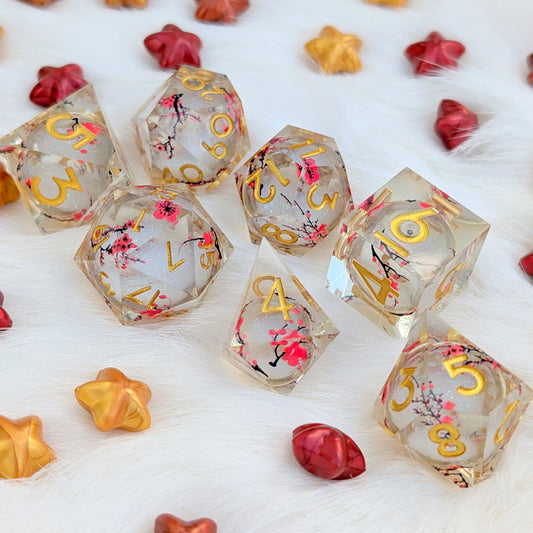 A set of transparent acrylic polyhedral dice with gold numbers and pink cherry blossom floral details, arranged on a white faux fur surface with decorative red and gold star-shaped props.