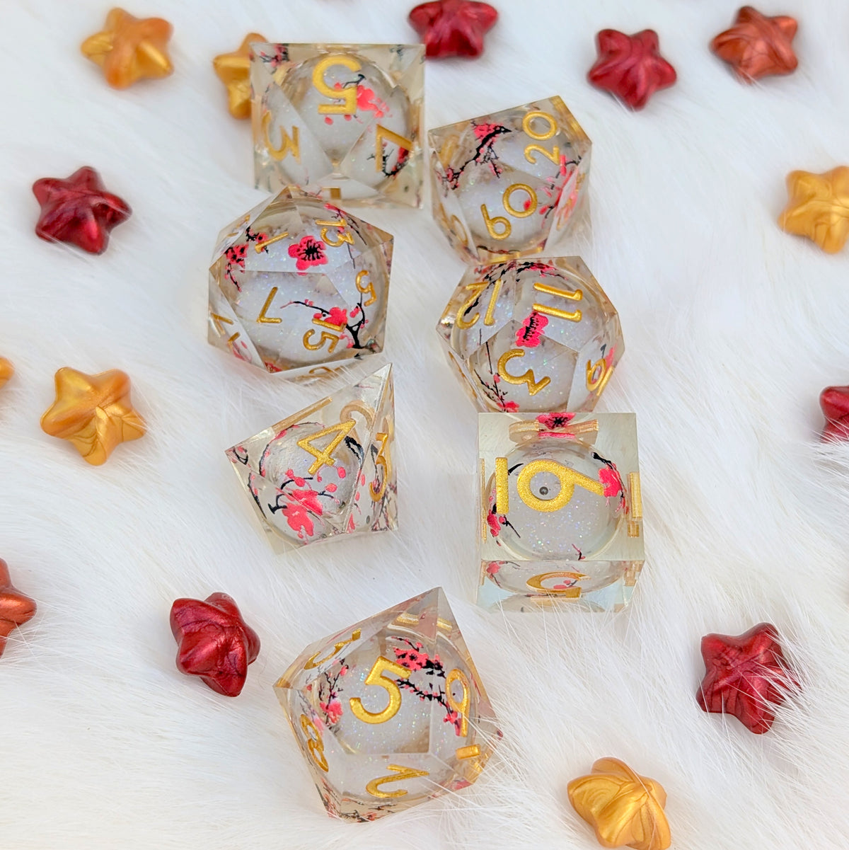 A set of transparent acrylic polyhedral gaming dice with gold numbers and pink cherry blossom designs, arranged on a white faux fur background with decorative red and gold star-shaped props.