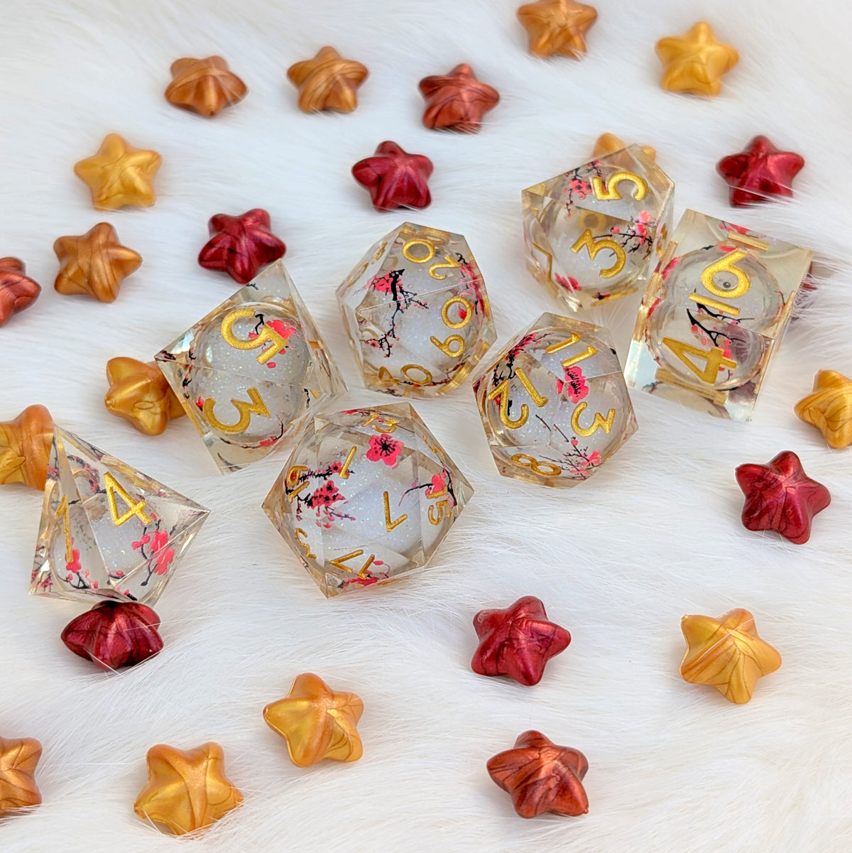 A set of clear resin polyhedral gaming dice with gold lettering and pink cherry blossom floral details, arranged on a white faux fur surface alongside decorative metallic star-shaped beads in gold, orange, and red.