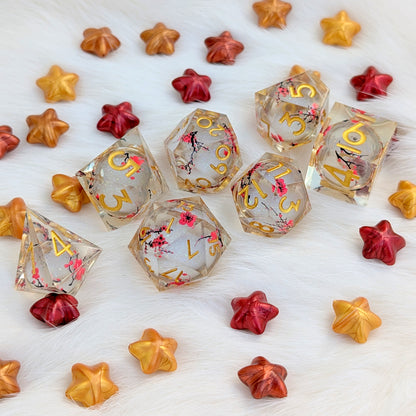 A set of clear resin polyhedral gaming dice with gold lettering and pink cherry blossom floral details, arranged on a white faux fur surface alongside decorative metallic star-shaped beads in gold, orange, and red.