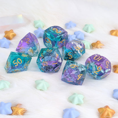 A set of translucent polyhedral gaming dice with purple, blue, and teal iridescent coloring and gold numbering, arranged on a white faux-fur surface with pastel star-shaped props.